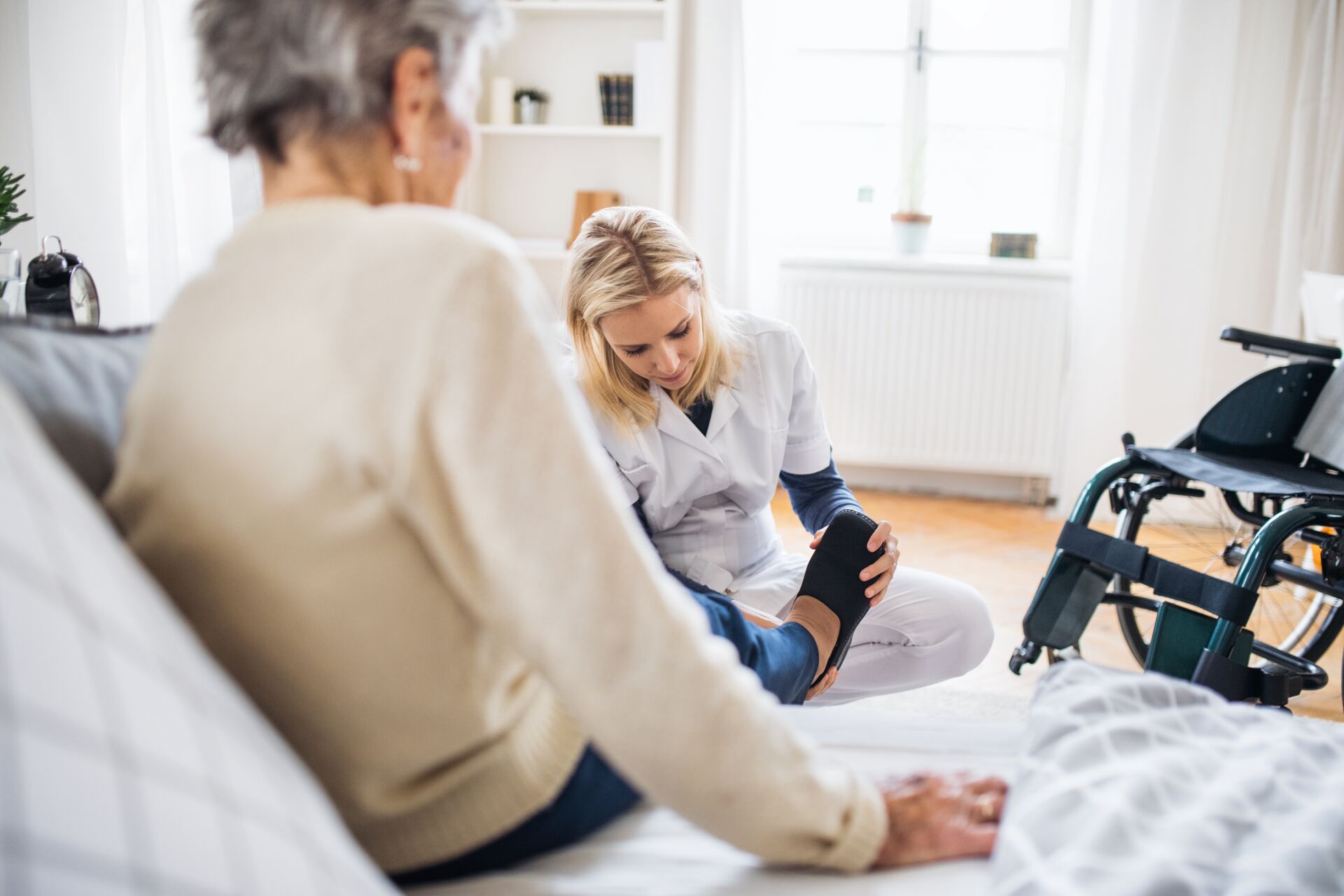 Eine blonde Pflegekraft hilft einer älteren Frau, die auf einem Sofa sitzt beim Schuhe anziehen. Im Hintergrund steht ein Rollstuhl.