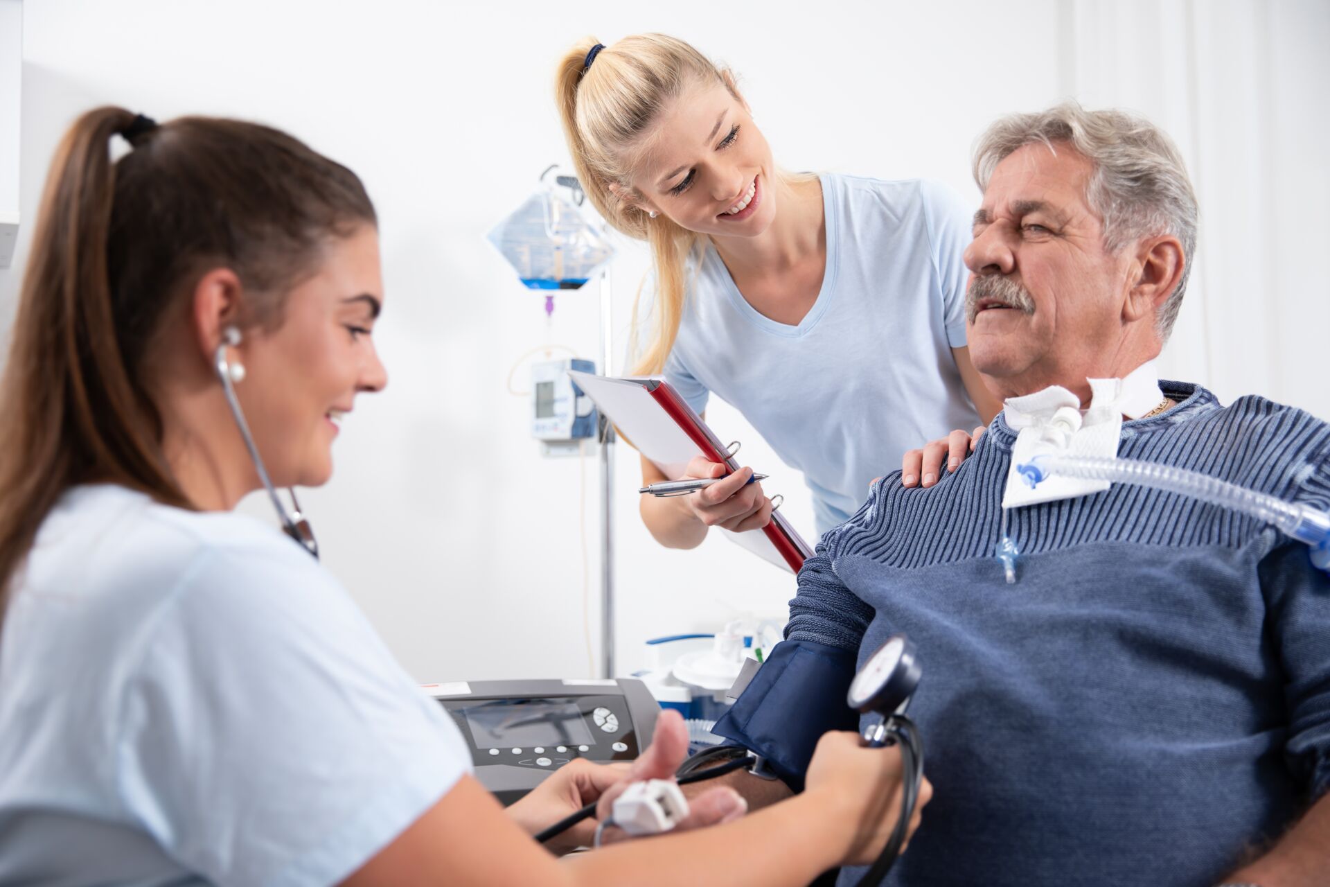 Eine junge Krankenschwester misst den Blutdruck eines älteren Herrn, während eine zweite Krankenschwester lächelnd daneben steht.