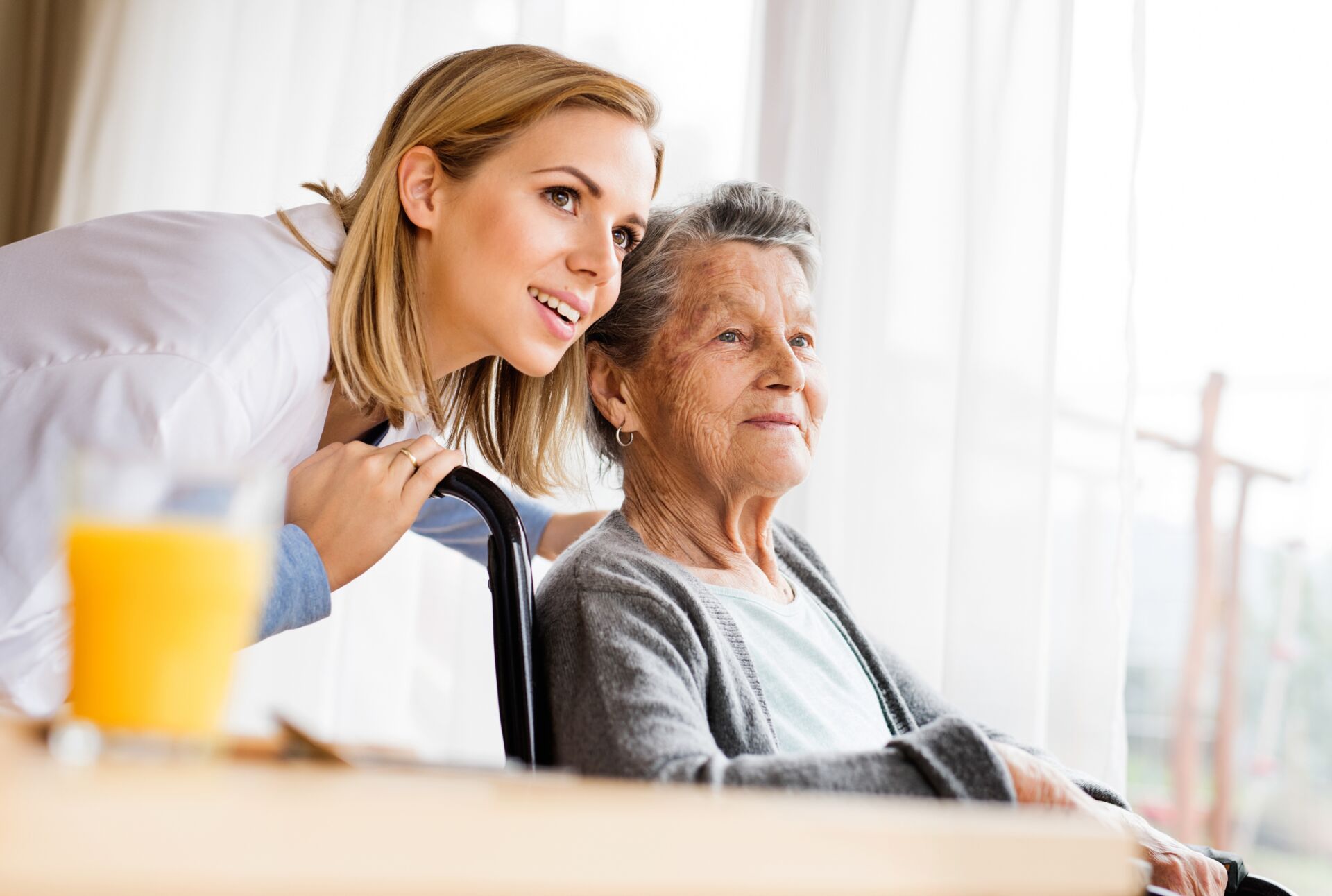 Eine Krankenschwester schaut mit einer älteren Frau, die im Rollstuhl sitzt aus dem Fenster